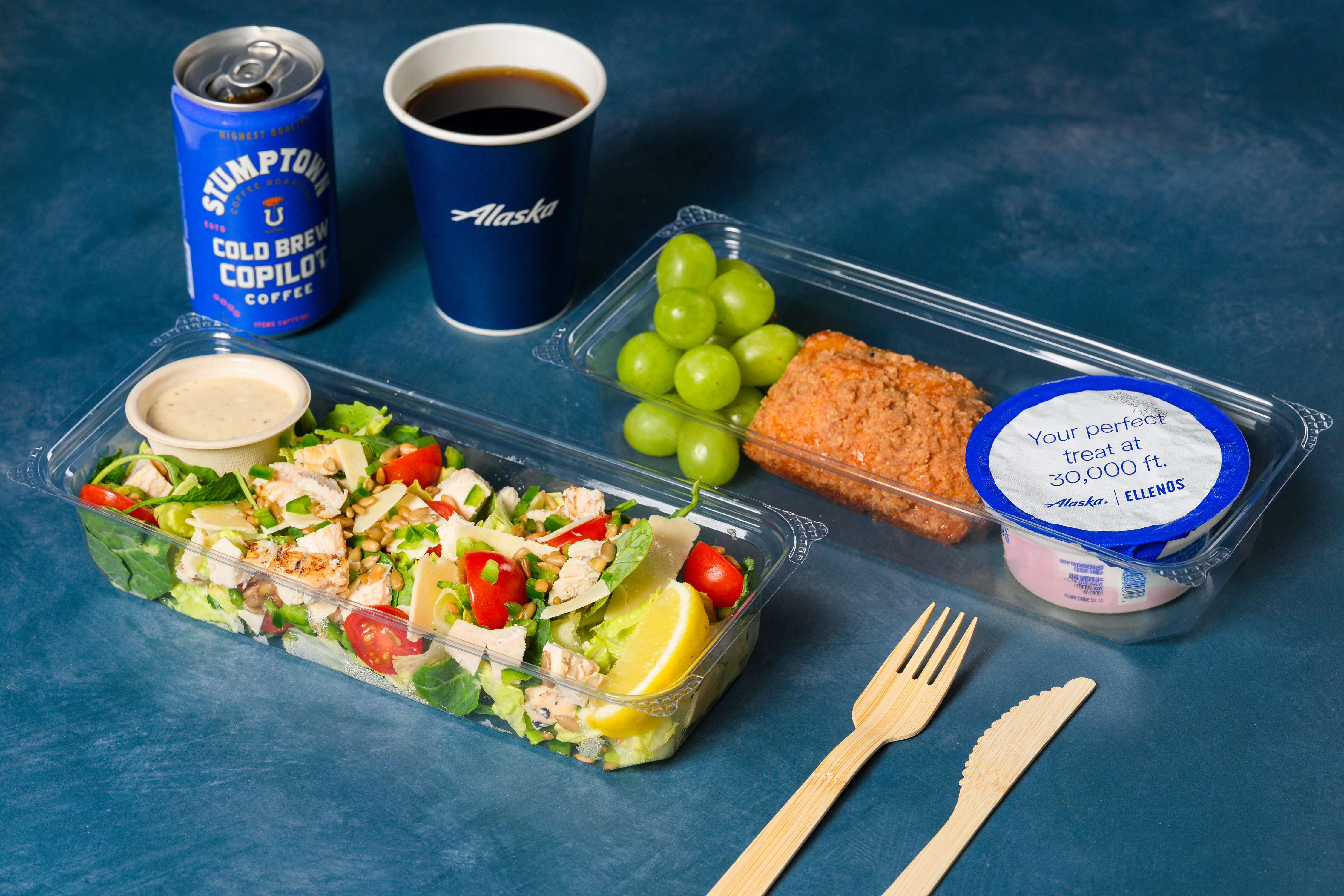 Alaska Airlines Main Cabin salad and an Ellenos yogurt breakfast plate with grapes and tea loaf, alongside hot coffee and Stumptown cold brew.