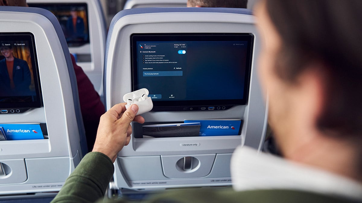 Passenger holding wireless earbuds beside an American Airlines seatback screen displaying the Bluetooth pairing menu.