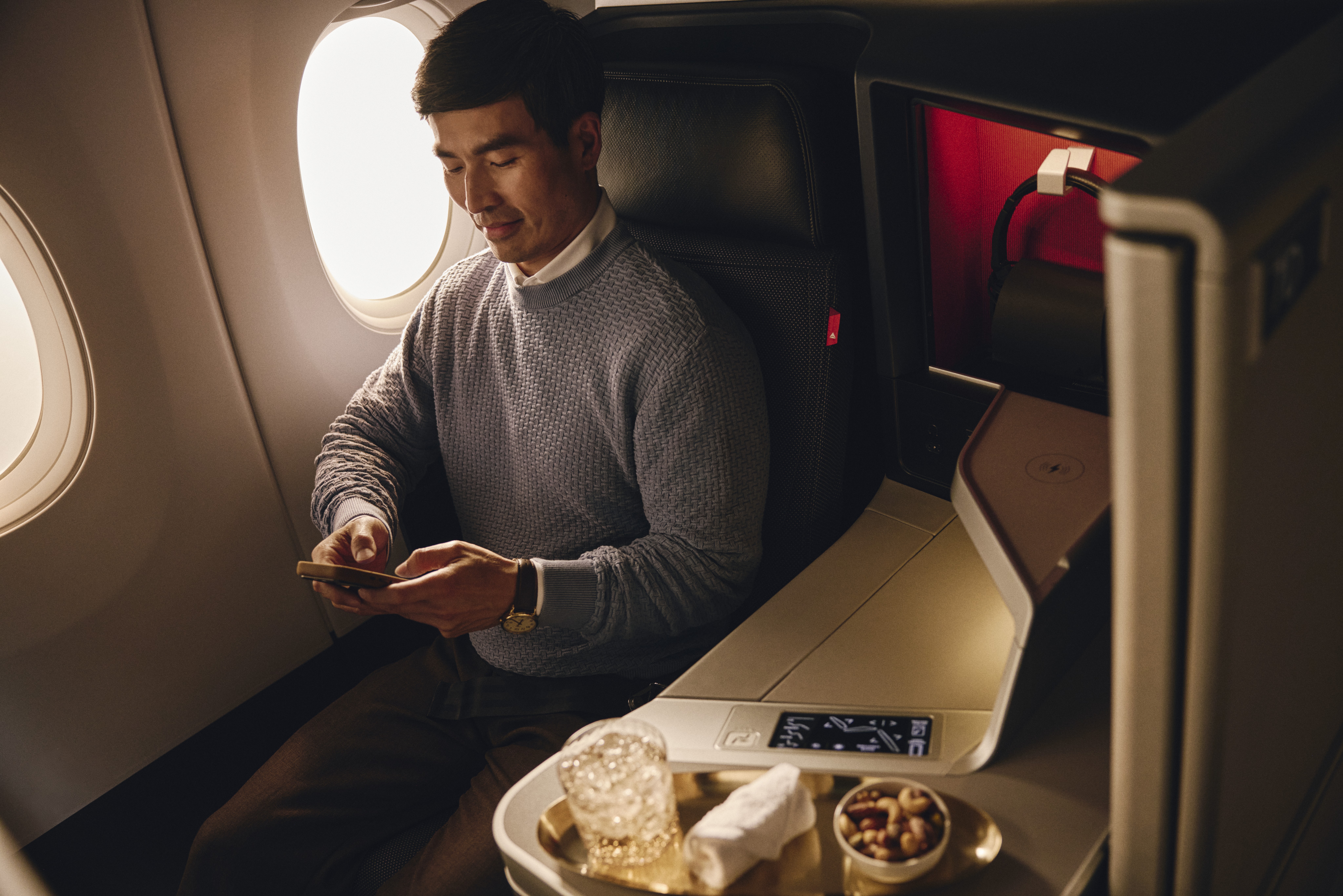 Passenger seated by the window in a Delta One suite, with a drink, towel, and nuts on the side console.